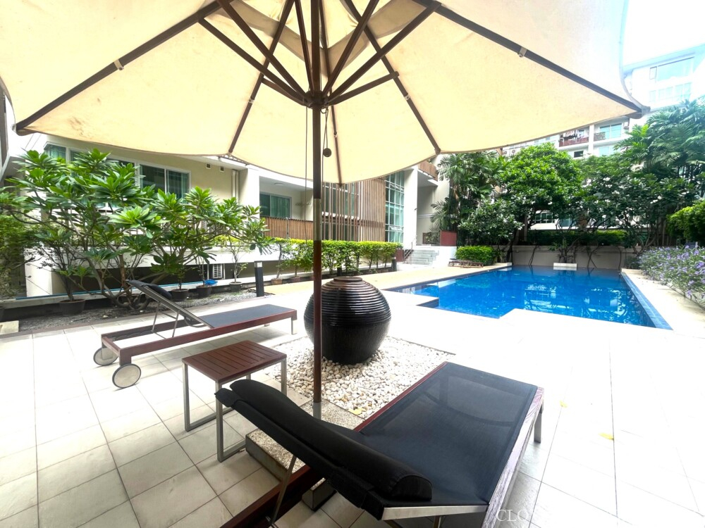 Poolside relaxation area at The Clover Thonglor. Features dark sun loungers, umbrella, tiled deck, and a contemporary urn accent next to the blue pool.