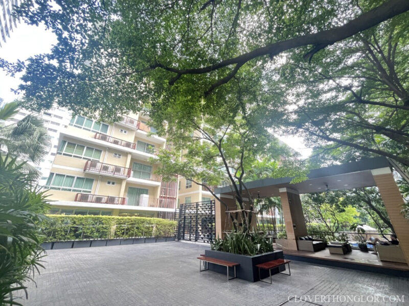Courtyard in front of pavilion Central courtyard at The Clover Thonglor, showing the residential building exterior, a paved gathering area, and the shaded outdoor relaxation pavilion.