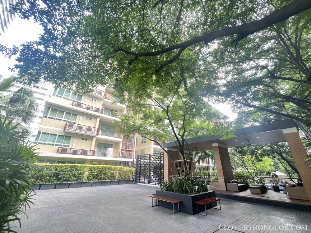 Central courtyard at The Clover Thonglor, showing the residential building exterior, a paved gathering area, and the shaded outdoor relaxation pavilion.