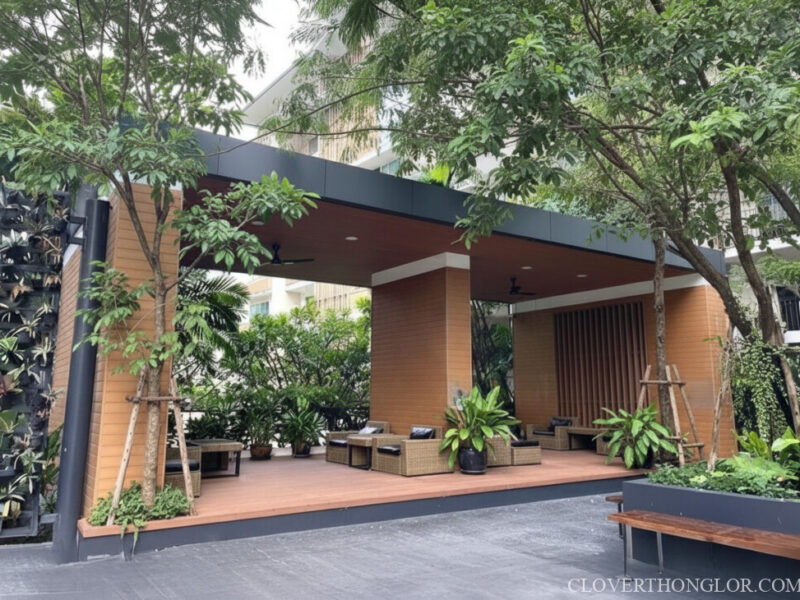 Stylish outdoor relaxation pavilion at The Clover Thonglor. Features a wood deck, comfortable rattan seating, and modern green vertical wall garden.