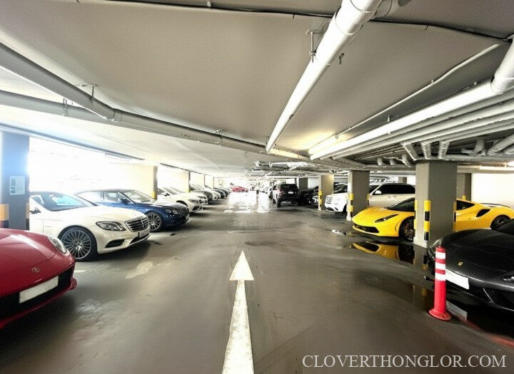 Secure underground car park at The Clover Thonglor. Features rows of parked vehicles and a painted directional arrow, offering ample parking space.