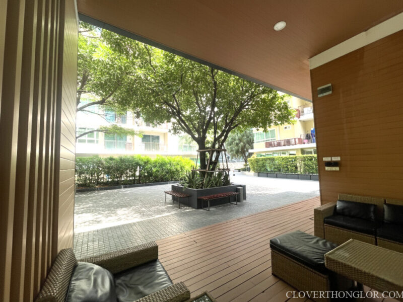 Shaded outdoor lounge area at The Clover Thonglor, with rattan seating on a wood deck, overlooking a central courtyard with a mature tree.