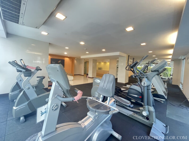 Close-up of the cardio equipment in The Clover Thonglor's gym, showing stationary bikes and ellipticals. The bright facility leads into a common corridor.