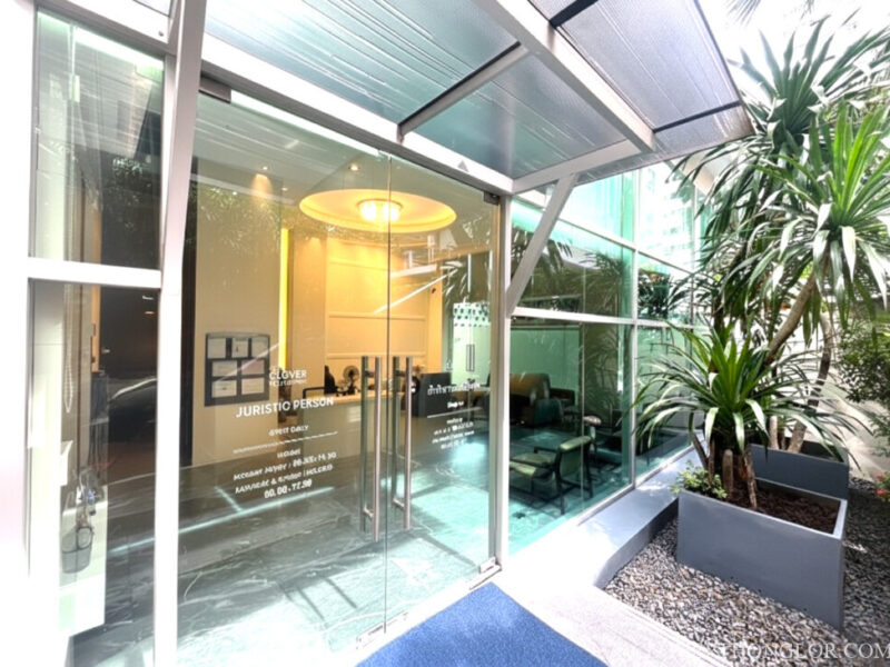 Modern glass entrance to the Juristic Person office at The Clover Thonglor Residence, highlighted by bright interior lighting and green plants.