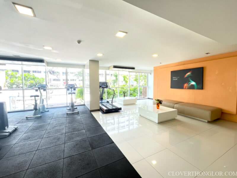 Modern multi-functional gym space at The Clover Thonglor. Features cardio equipment, a bright white tiled lounge area, and a large wall-mounted screen