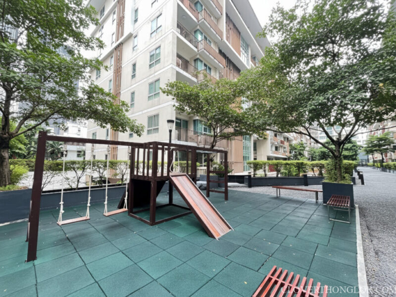 Children's play area at The Clover Thonglor, featuring a wooden slide and swing set on a safe green rubber surface, shaded by surrounding trees.