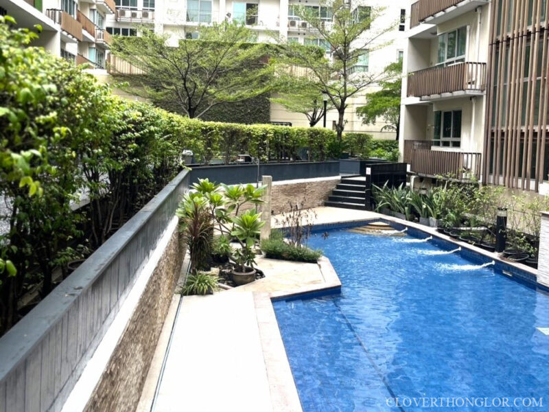 Corner view of the Clover Thonglor's swimming pool, showing a tiled edge, small water features, and steps leading to a raised, private garden deck.