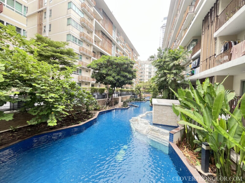 A very long, winding swimming pool at The Clover Thonglor, surrounded by dense tropical landscaping, nestled between the low-rise condo buildings.