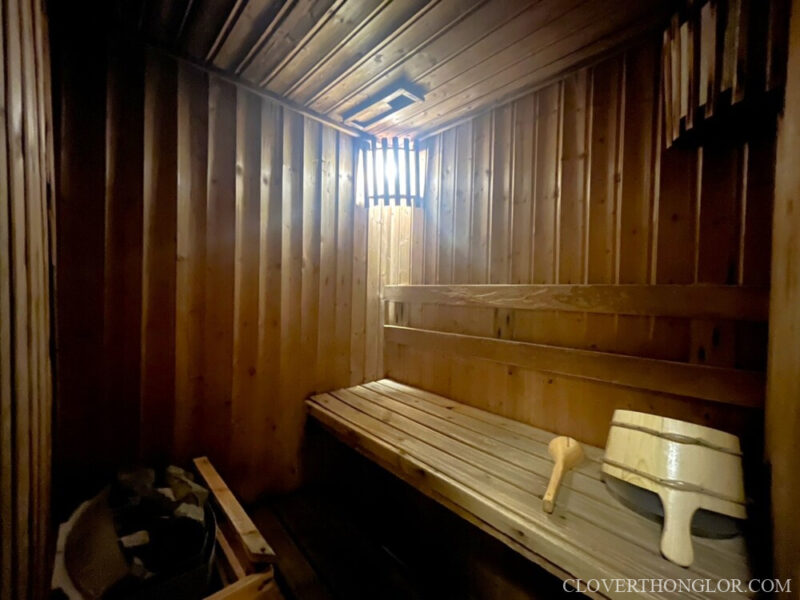 Interior of the warm, traditional sauna room at The Clover Thonglor's gym, featuring multi-level wooden benches, heat rocks, and a ladle/bucket set.