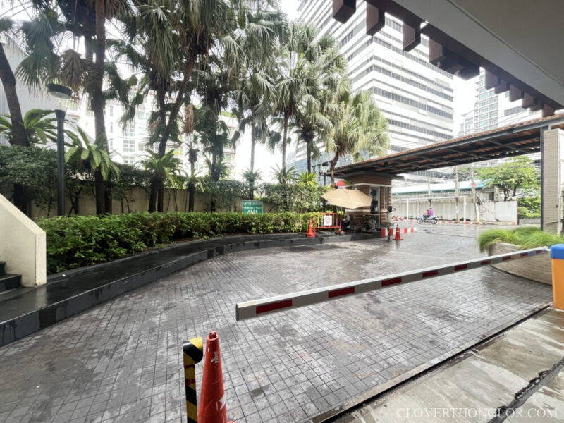 Main vehicle entry point and security gate barrier at The Clover Thonglor, featuring a paved driveway and tropical plants, with tall city buildings visible.