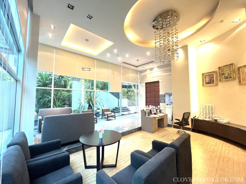 sofa in the lobby with Spacious, sunlit lobby lounge and reception at The Clover Thonglor. Features dark seating, a chandelier, and glass walls opening to the garden.