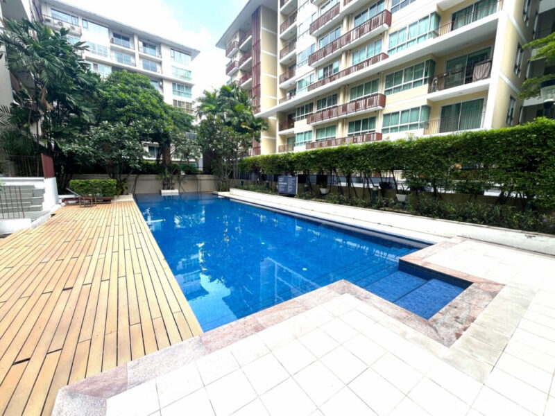 swimming pool perspective Rectangular lap swimming pool at The Clover Thonglor. Features a wooden sun deck, stone border, and a hedge separating it from the low-rise condos.