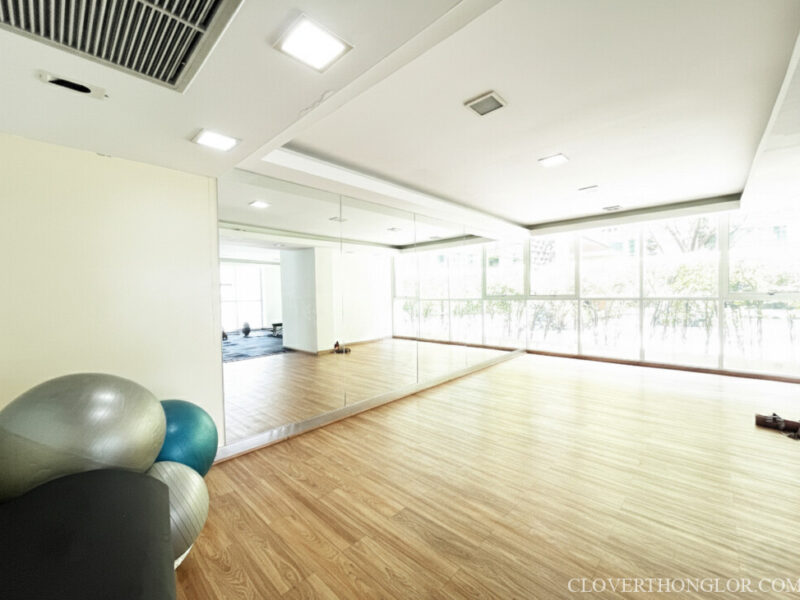 Sunlit yoga and stretching studio in the Clover Thonglor gym. Features warm wood floor, full-height mirrored wall, and large windows with natural light.